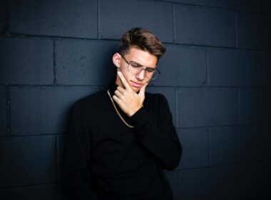 man leaning on wall while looking down wearing eyeglasses and necklace with right hand on chin